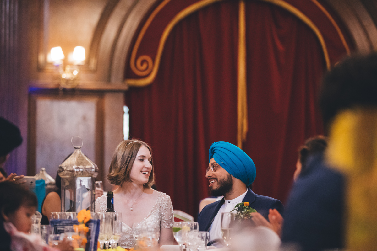 Bride and Groom seated looking at each other smiling at their wedding reception in the Main Hall at Porchester Hall London. There is a large red curtain with yellow trim in the background. The groom is wearing a blue turban and glasses
