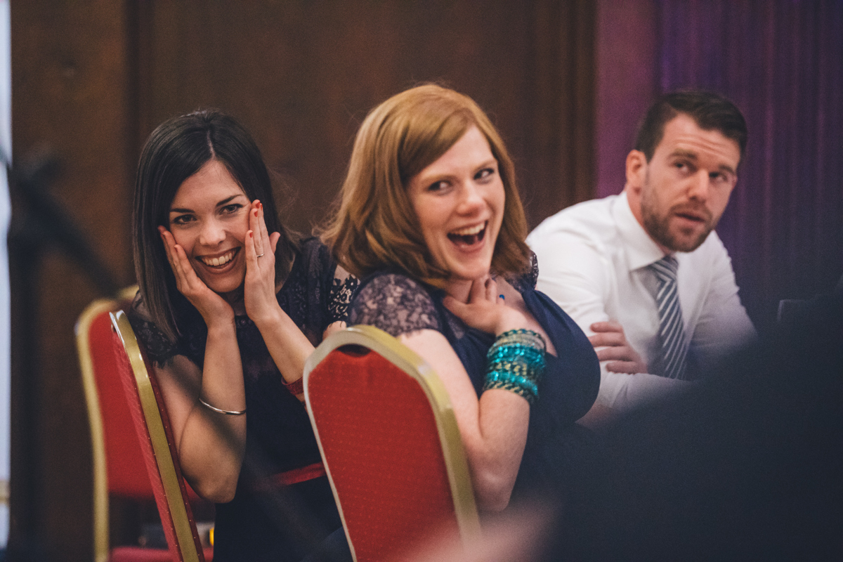 Two women sat on red chairs, both wearing dark blue dresses, looking shocked and smiling during speeces at a wedding at Porchester Hall, London, with a man in a white shirt and striped tie sat in the background