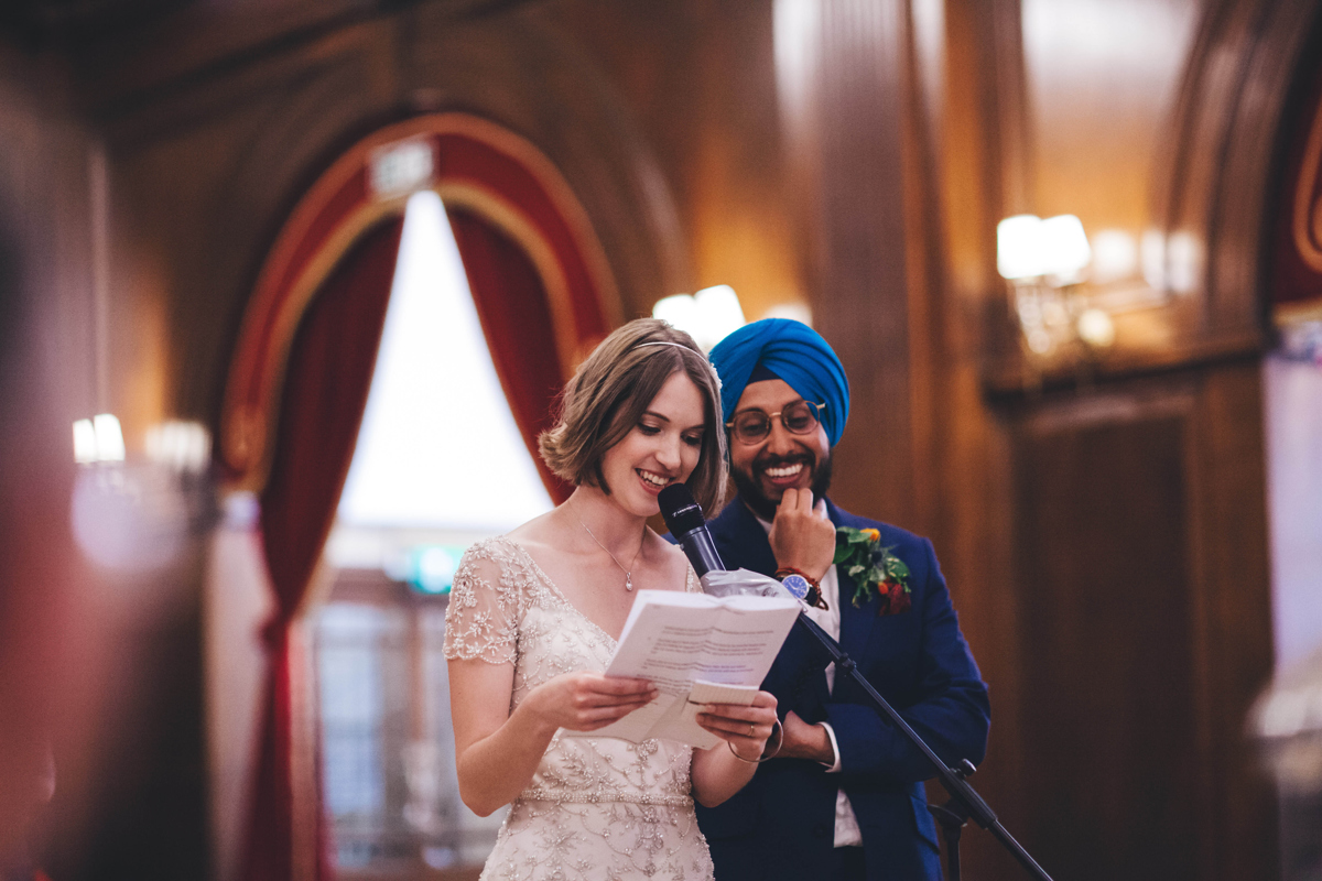 Bride and groom stood in front of a microphone reading from a piece of paper, hled by the bride, at a wedding reception at Porchester Hall, London