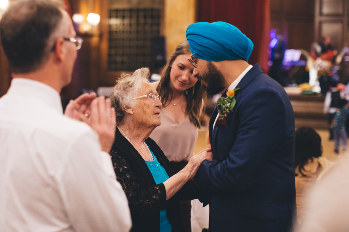 Groom, wearing a turban and glasses, holding the hands of a old lady, also wearing glasses, with a young woman in the background and a middle-aged man in the foreground with his back to the camera