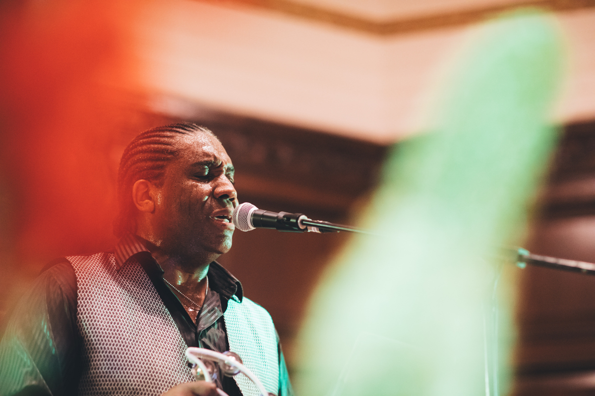 A black man with braids in his hair singing into a microphone and holding a tambourine at Porchester Hall, London