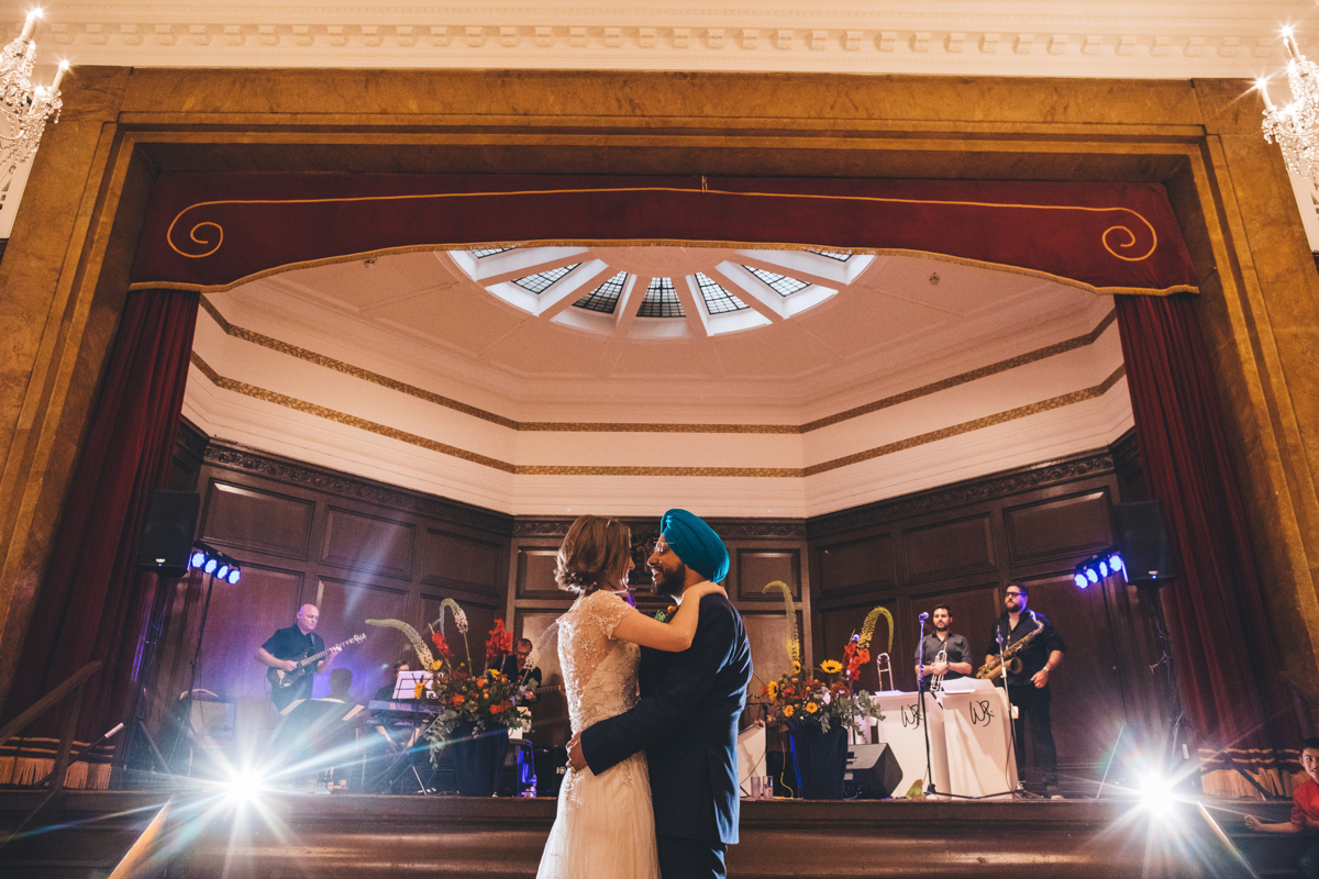 Bride and Groom with their arms around each other during their first dance at Porchester Hall, London. There is a band in the background on a wood panelled stage with a large sky light above