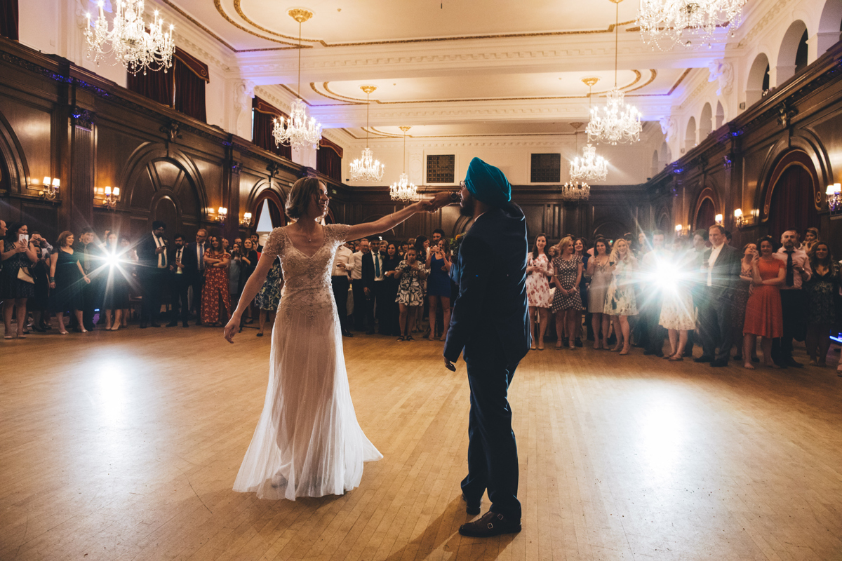 Bride and Groom on the dancefloor during their firt dance in the Main Hall at Porchester Hall, London. The wedding party are stood in the background watching the couple with many taking photographs. The bride has her left arm and hand outstretched which the groom is holding with his right hand