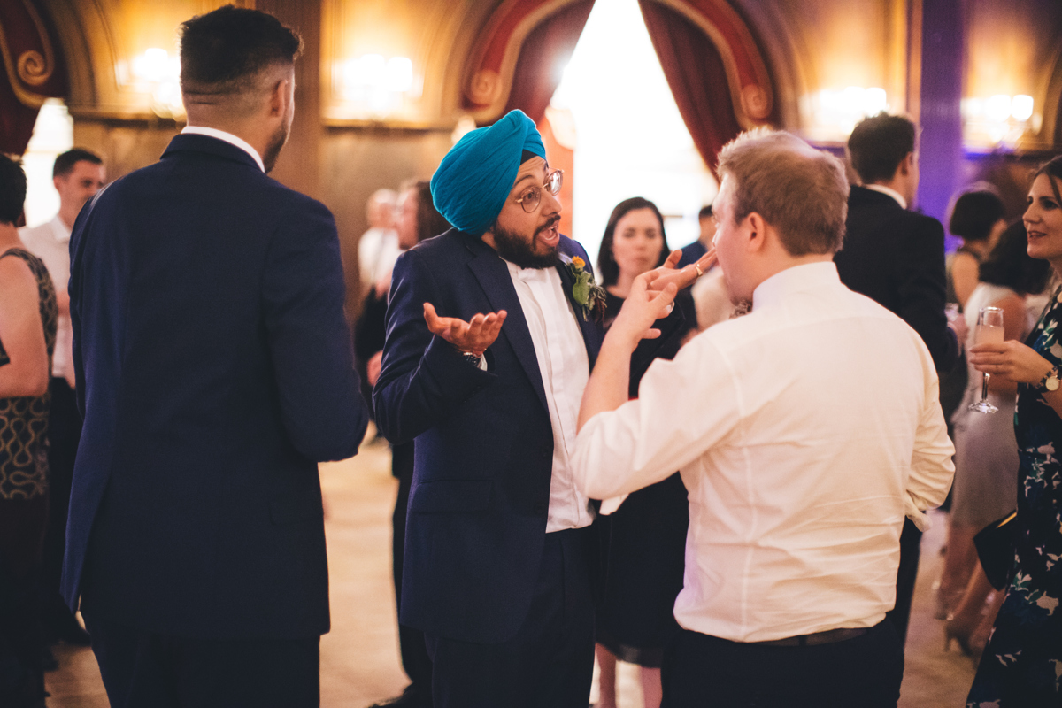Groom, wearing a blue turban, talking to a male in a white shirt. The groom is holding both hands upwards with a surprised look on his face