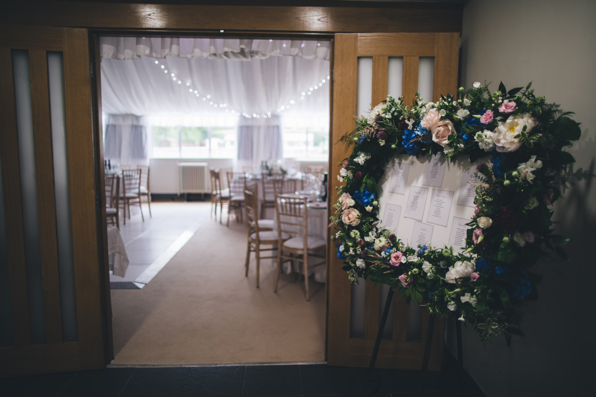 A table plan mounted on a board surrounded by flowers to the right of a doorway which looks into a room set out for a wedding reception at Bardsey Village Hall