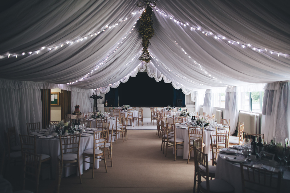 Village Hall dressed ready for a wedding reception with white material on the ceiling lined with white fairy lights. There are circular tables surrounded by wooden chairs