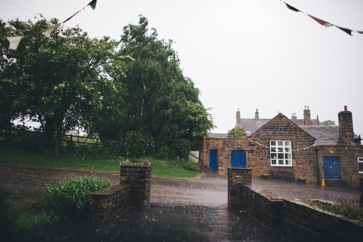 Rain pouring down on the road in Bardsey village, Leeds with a stone building with bunting on the other side of the road with green trees to the left of the picture