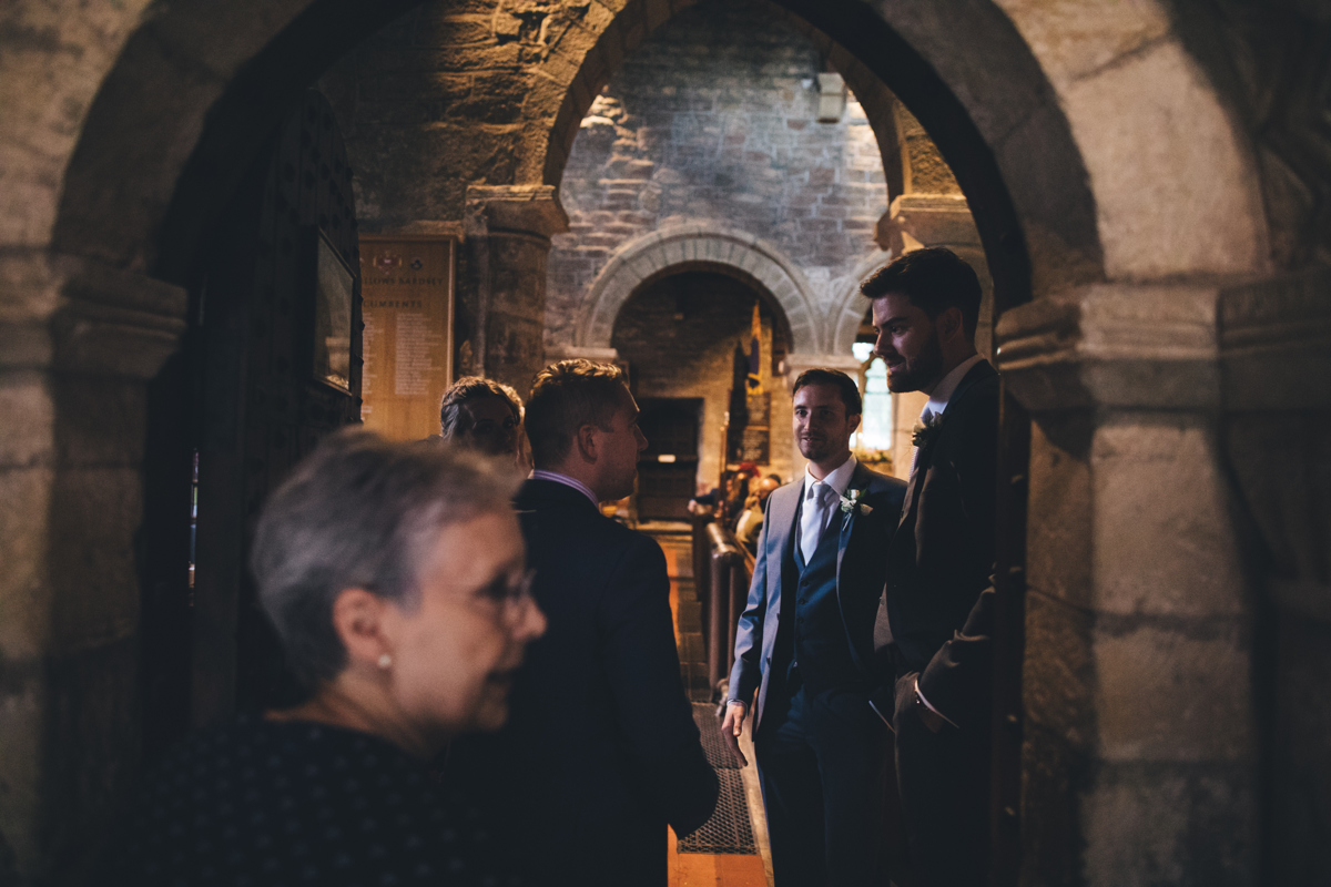 View from the doorway at the church in Bardsey village, Leeds, looking into the church where there are three men wearing suits talking to each other with an older lady in the foreground looking to the right