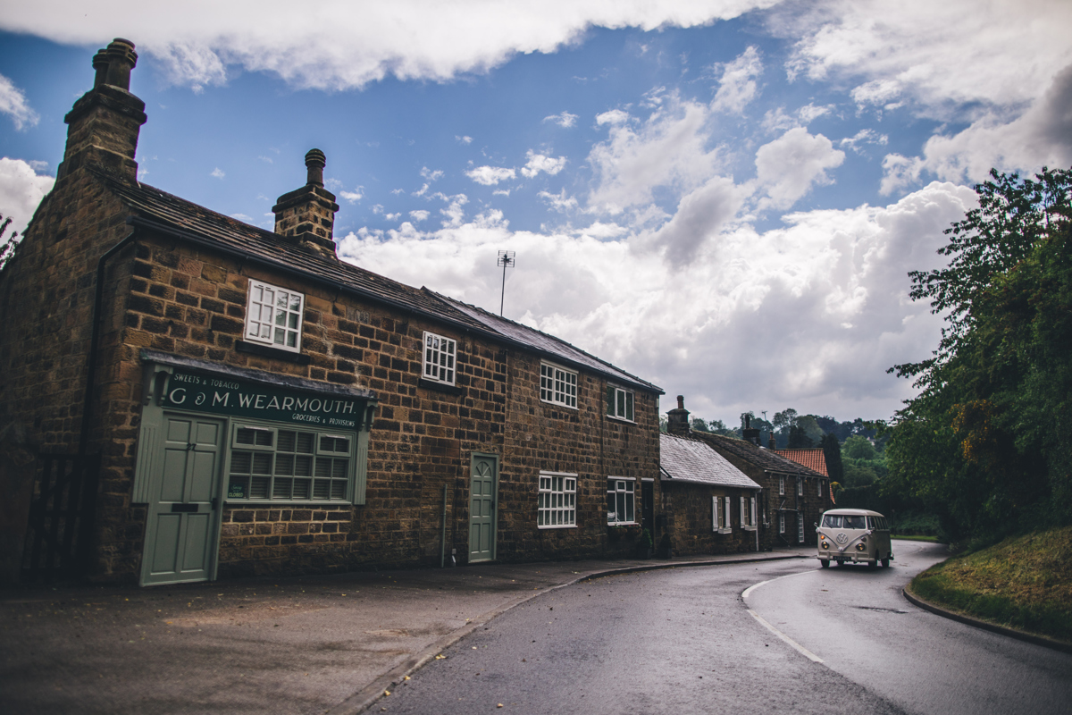 View of stone buildings to the left of the picture and a vintage white VW Campervan driving up the road on the right of the picture with blue skies and white clouds in the background