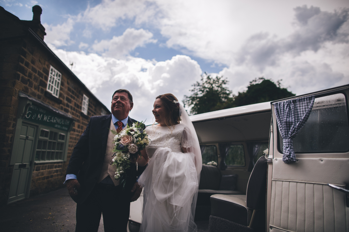 Bride and her father stood next to a white VW Campervan. The bride is holding a bouquet of flowers