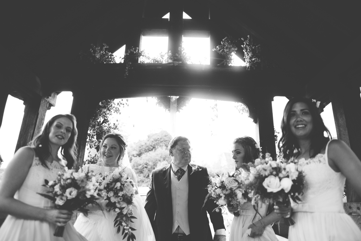 Black and white photograph of father of the bride in the centre with his daughter and three bridesmaids either side of hi under a wooden archway surrounded by flowers