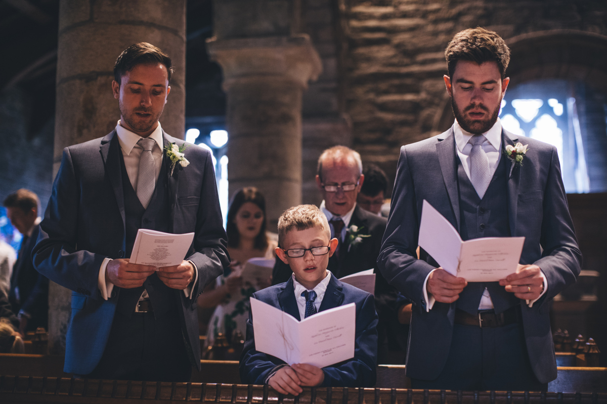 Two groomsmen either side of a young boy, all of them are stood up, singing from an order of service which they are holding in their hands