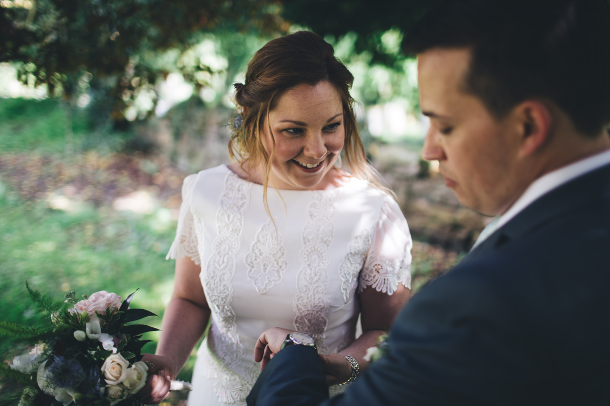 Bride holding a bouquet of flowers smiling towards her husband who is looking down at his watch