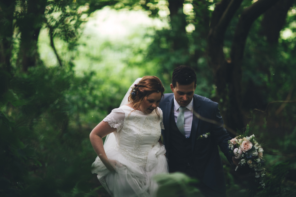 Bride and groom walking through woodland. The bride is holding up the bottom of her dress and the groom is holding a bouquet of flowers