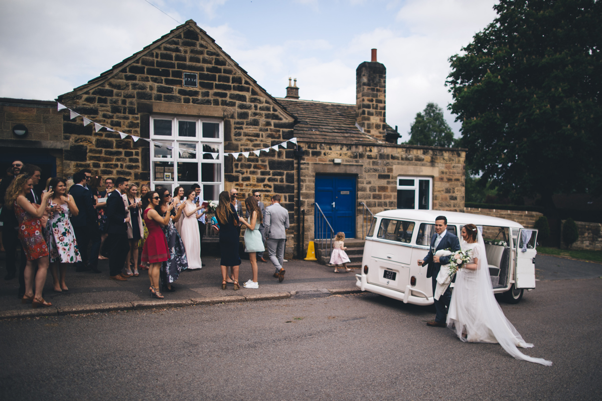 Bride and groom walking from a white VW campervan towards the wedding party who are stood in front of a stone building with bunting across the window in Bardsey, Leeds