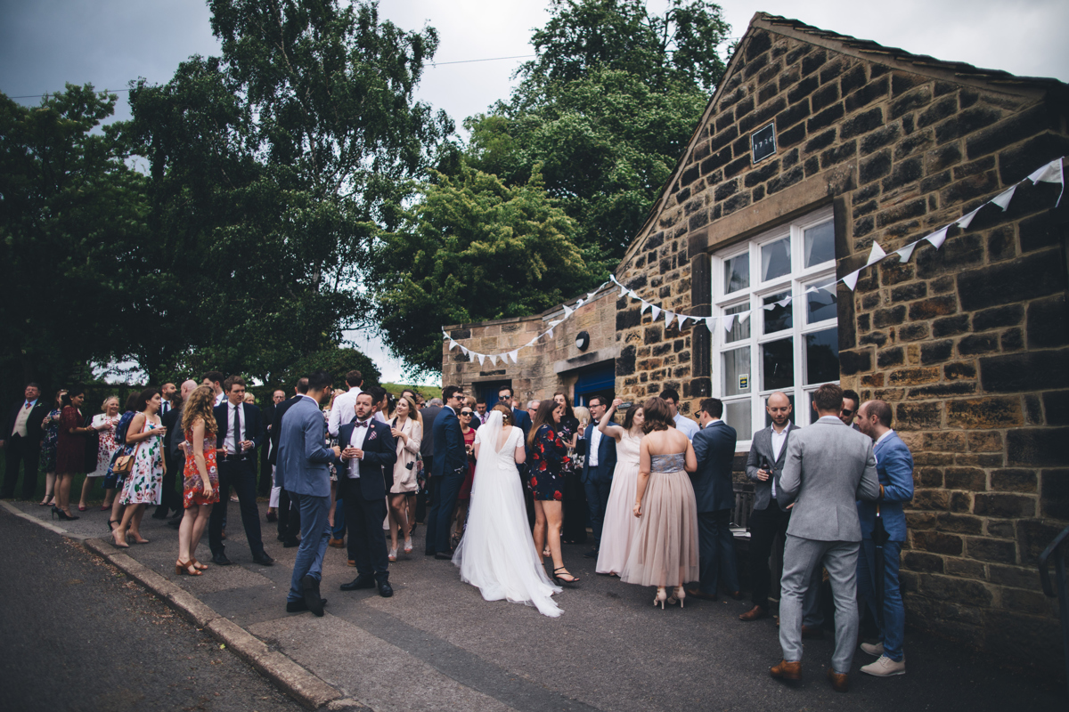 Wedding party stood chatting outside a stone building with bunting draped across the front in Bardsey Village, Leeds