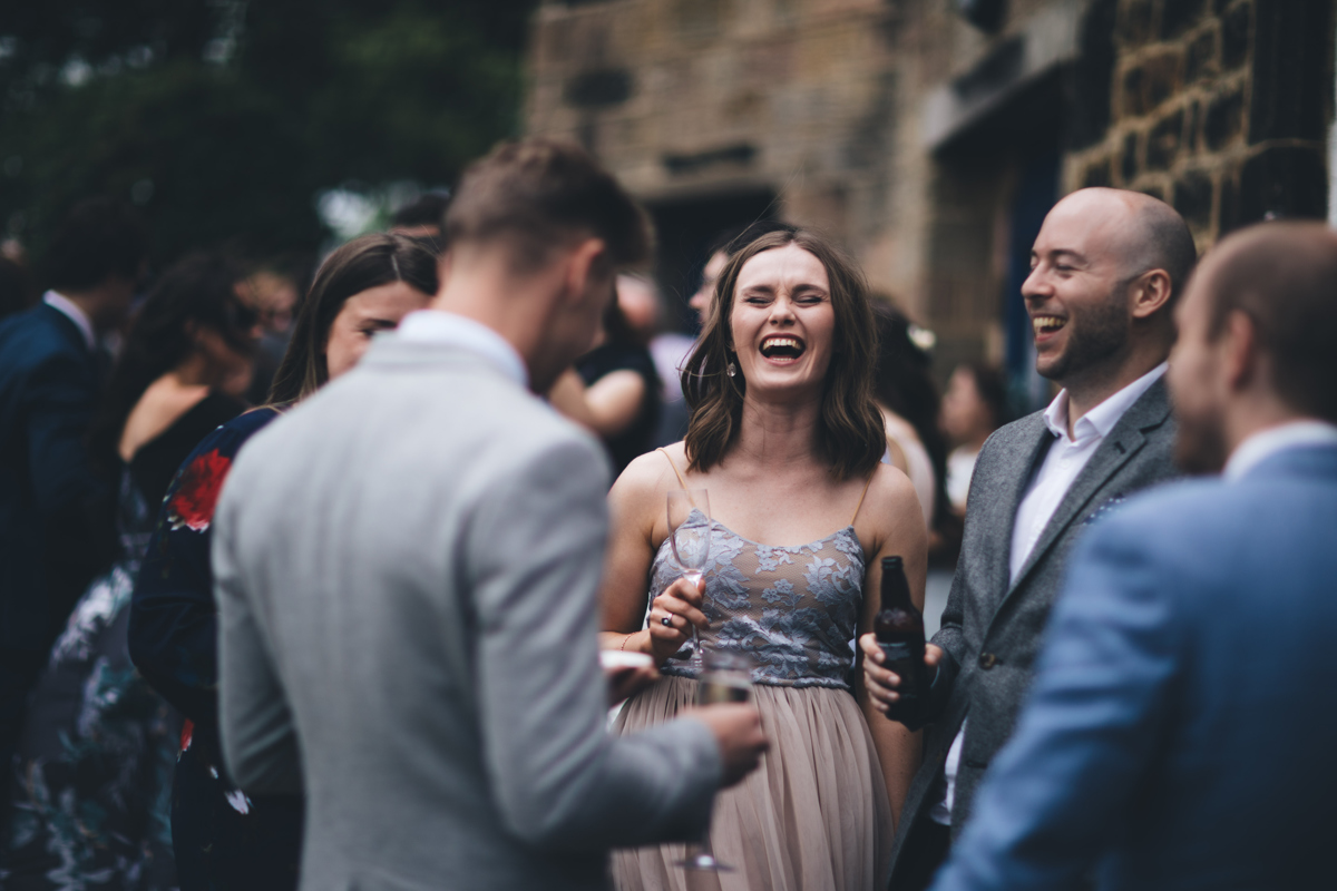 Female wedding guest laughijg with her mouth open surrounded by friends who are also laughing. They are all holding a drink in their hands
