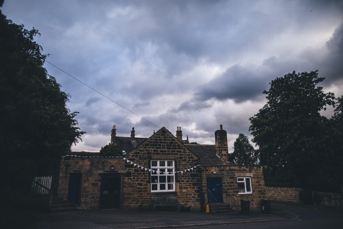 Stone building with blue doors and bunting across the window in Bardesy Village, Leeds. There are large trees either side of the building and a cloudy sky in the background