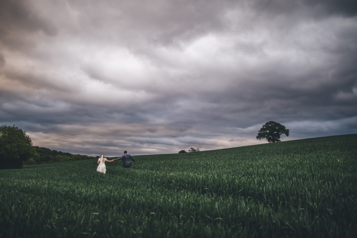 Bride and groom holding hands with outstretched arms walking through a green wheat field with a dark cloudy sky above them with trees in the background
