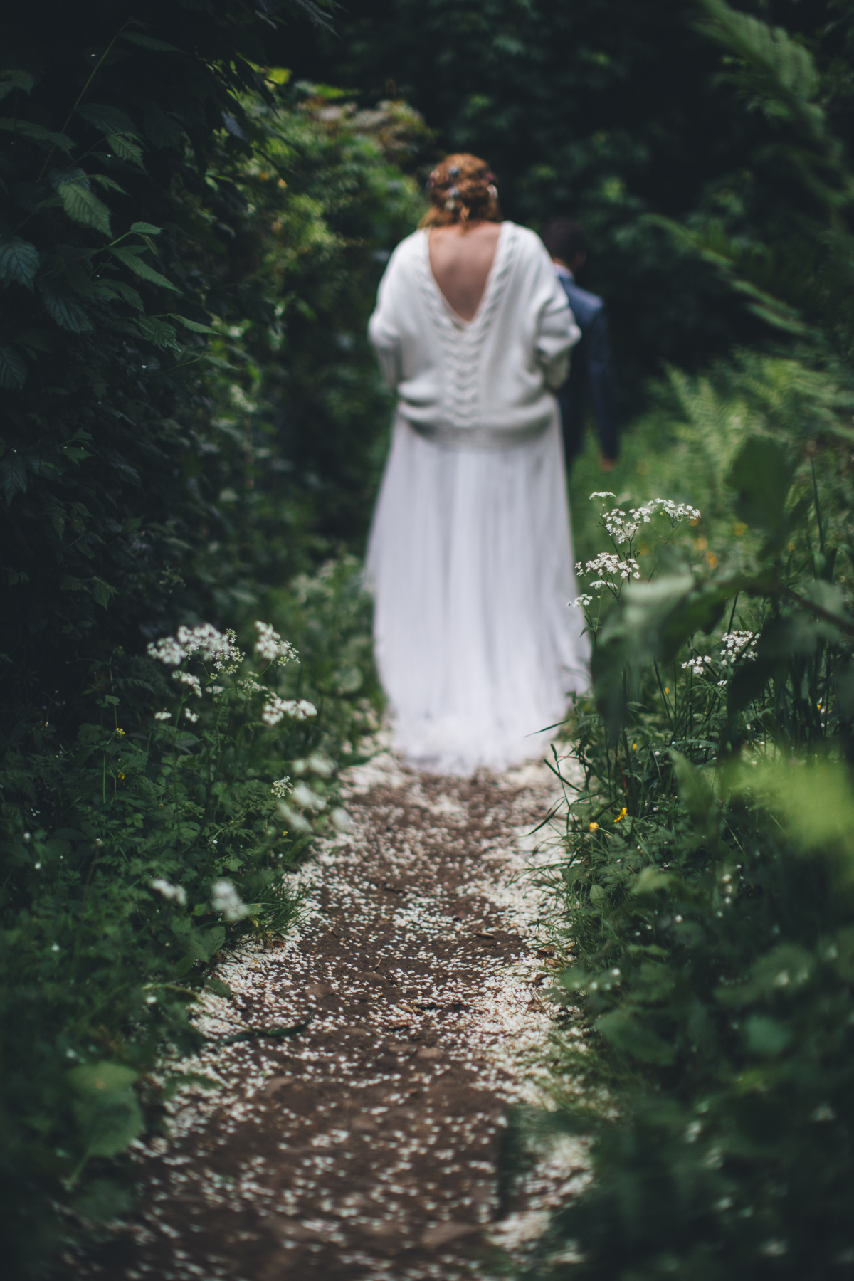 Bride walking away from the camera down a blossom lined path with green foliage surrounding her. The groom is walking in front of her and only his head and right arm can be seen. The bride is wearing a white jumper, with a low cut neckline, over her wedding dress