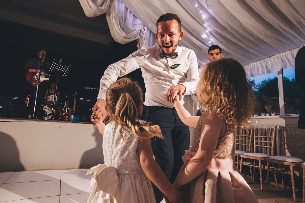 Man wearing a white shirt and a bowtie dancing with two young girls in dresses in the dancefloor at a wedding in Bardsey Village Hall, Leeds