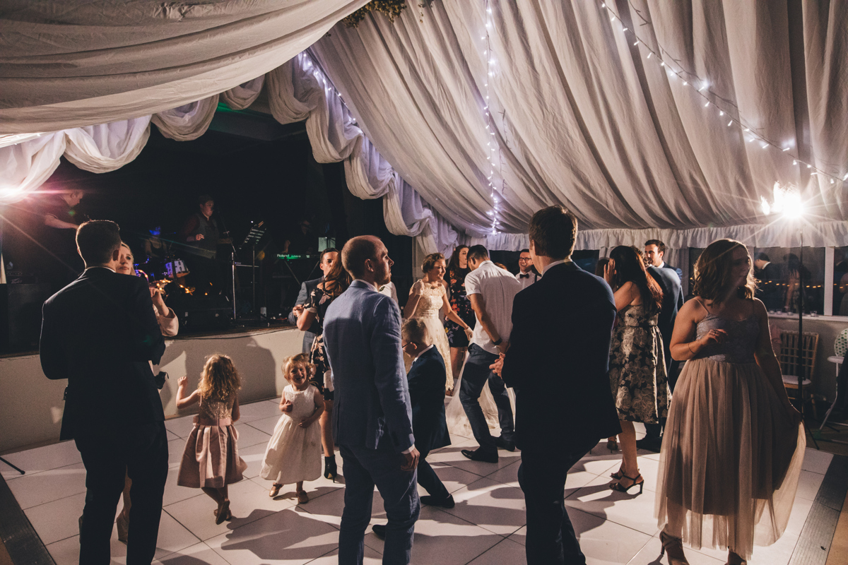 View of people dancing at a wedding at Bardsey Village Hall, Leeds. There is white material draped from the ceiling and fairy lights hanging across the material. There is a band playing in the background