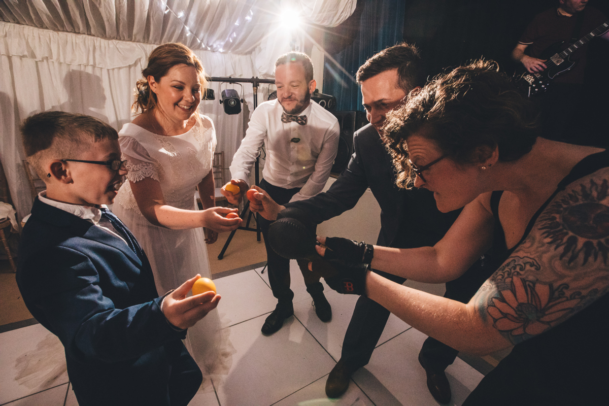 Bride and groom with two adults and a young boy on the dancefloor at bardsey Village Hall, Leeds, holding small yellow and orange balls. They are stood in a circle facing inwards holding their arms out