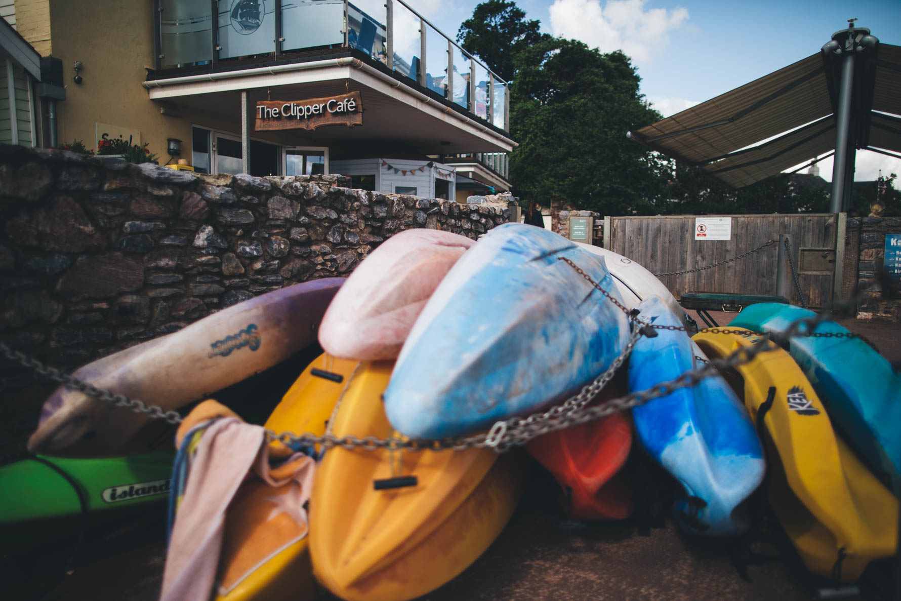 Colourful kayaks stacked up in front of a stone wall in front of The Clipper Cafe