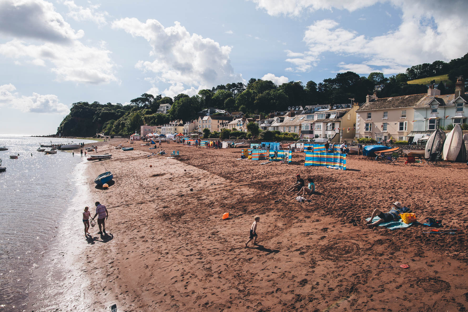 A beach next to a harbour in devon with pastel coloured houses overlooking the beach. There are a lot of windbreakers on the beach and a few people walking and sitting on the sand