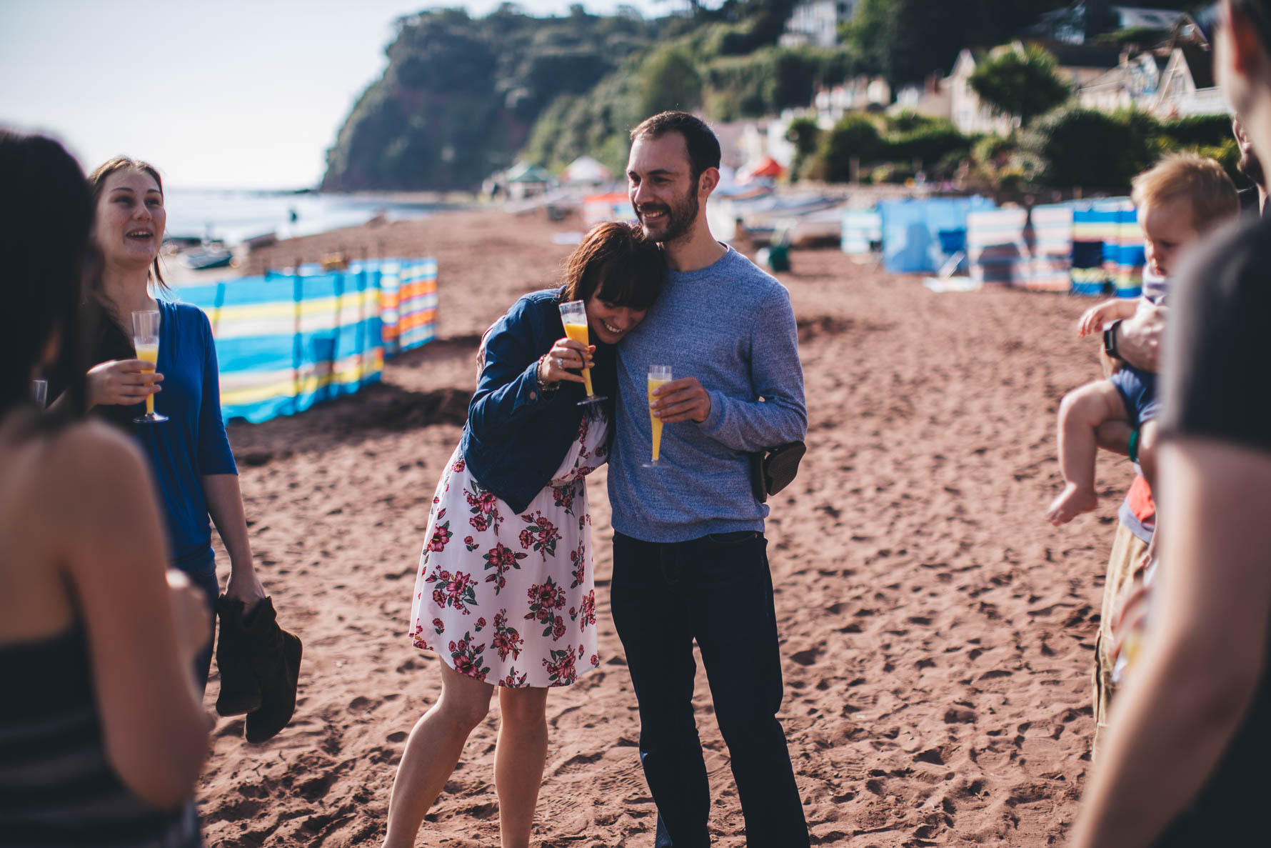 A man and a woman with their arms around each other stood on a beach in Devon holding glasses of bucks fizz in champagne flutes. There are other people on the edge of the picture also holding glasses of bucks fizz. There are lots of windbreakers on the sand in the background
