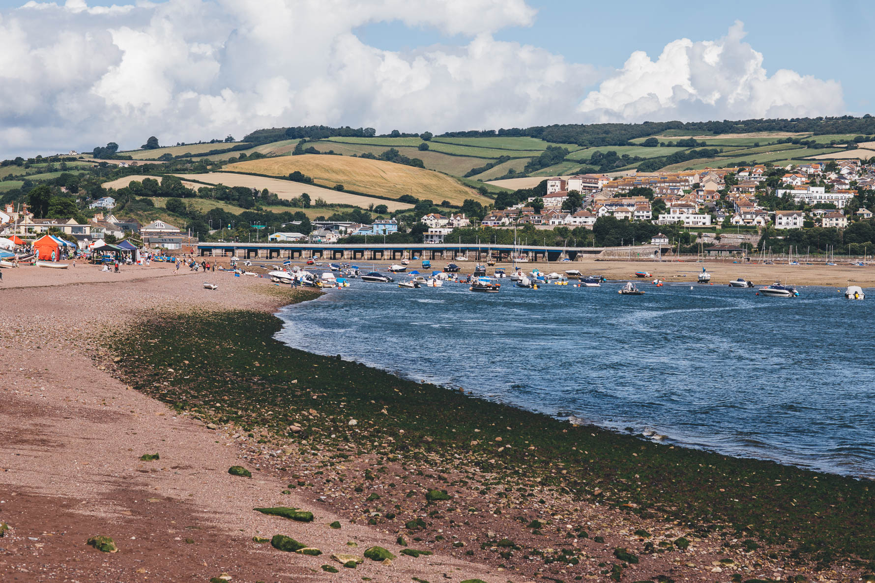 A scene of a beach and a harbour in Devon with houses and a patchwork of fields in the background below a blue sky filled with white clouds. There are lots of boats in the harbour