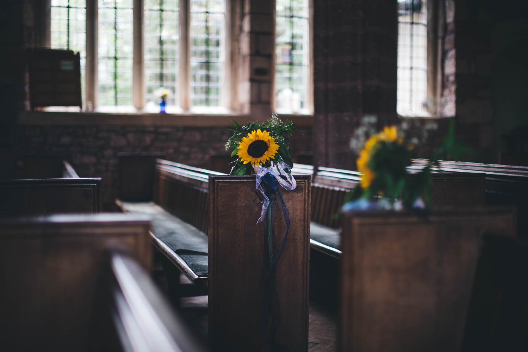 Picture of a flower arrangement with a large sunflower in the centre sat on the edge of a church pew. There are leaded glass windows in the background