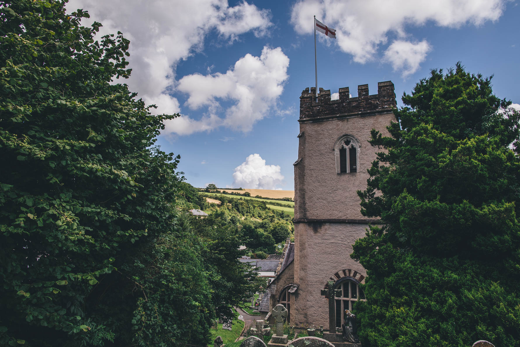 Castellated church with large trees to either side of it and a St George's cross flag flying from the top of the building against a blue sky with white clouds