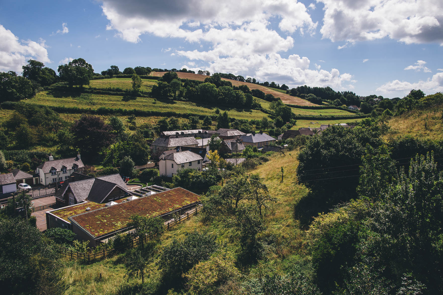 A rolling hillside village in Devonwith buildings in the centre of the picture and greenery in the foreground and background against a blue sky with white clouds