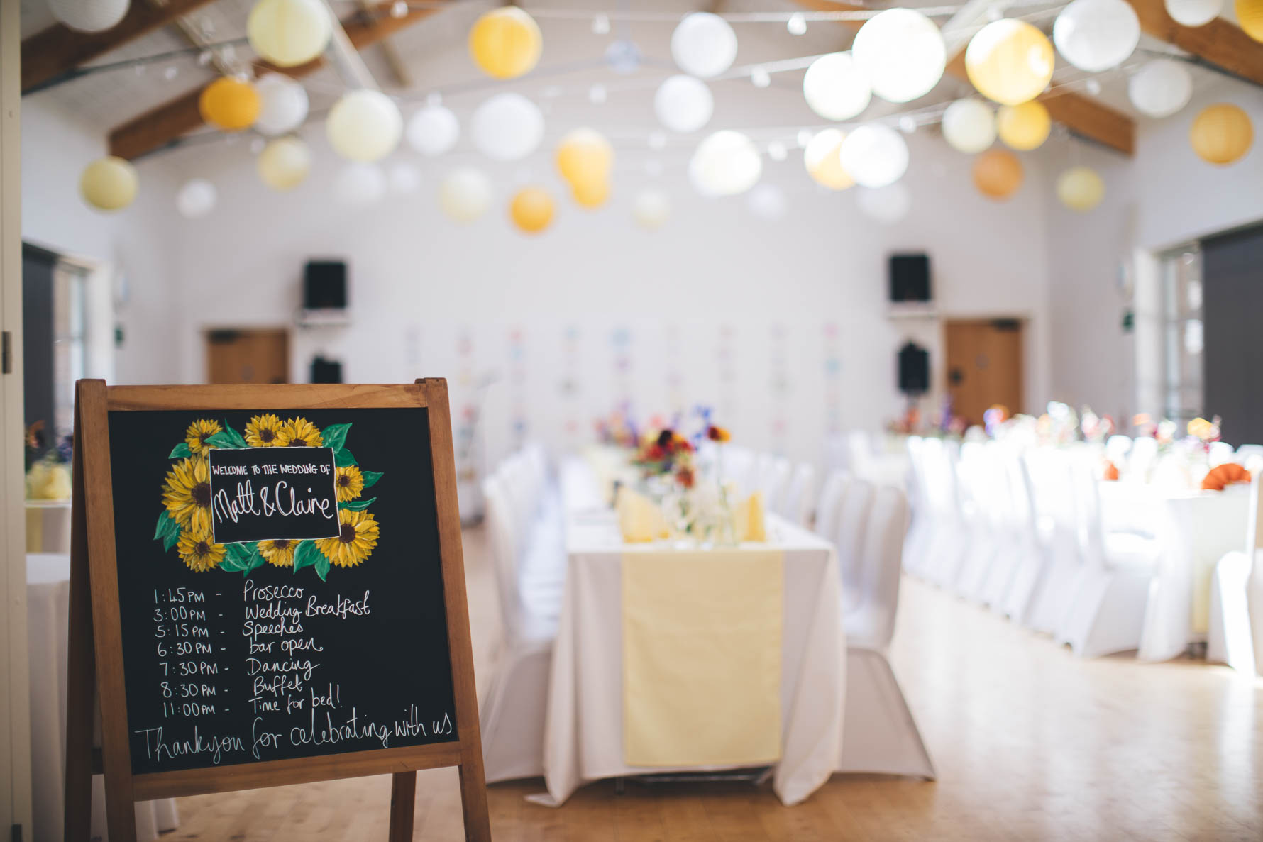 Village hall in Devon decorated for a wedding reception with white chair coverings and long white tables. There are white and yellow paper lanterns hung from the beams across the room and there is a chalkboard with a plan for the day's events in time order
