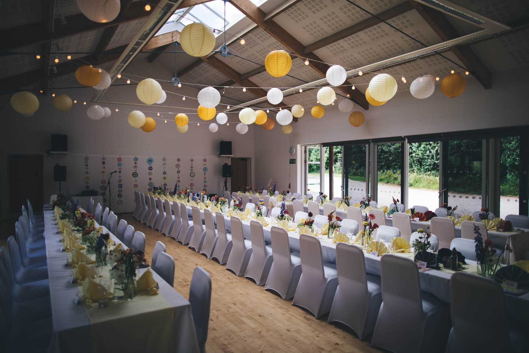 Village hall in Devon decorated for a wedding reception with white chair coverings and long white tables with floral arrangements along the tables. There are white and yellow paper lanterns hung from the beams across the room.