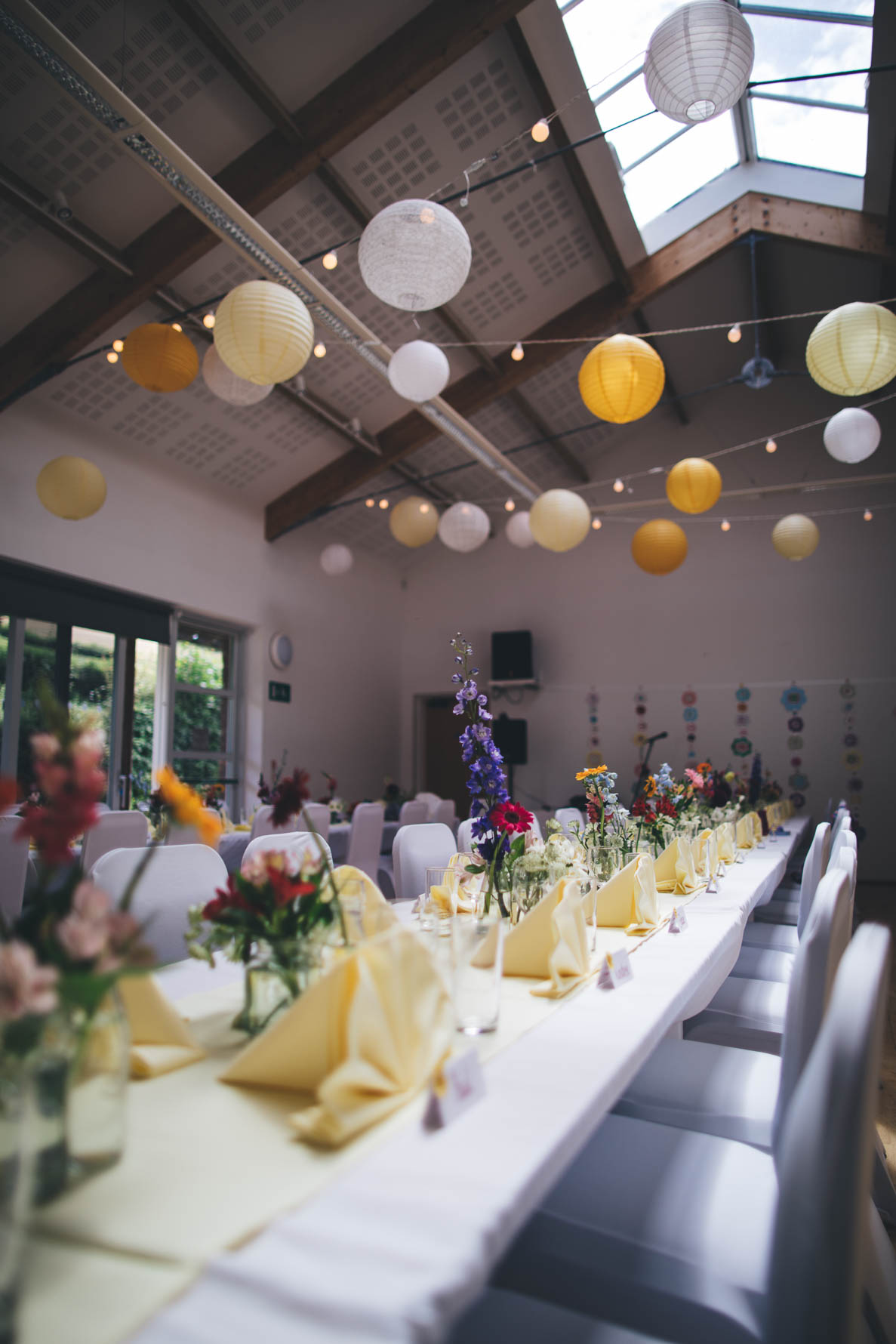 Village hall in Devon decorated for a wedding reception with white chair coverings and long white tables with floral arrangements along the tables. There are white and yellow paper lanterns hung from the beams across the room.