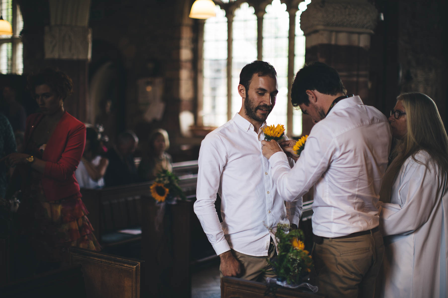 Best man pinning a sunflower onto the grooms shirt. They are stood inside a church and are both wearing white shirts and khaki chinos