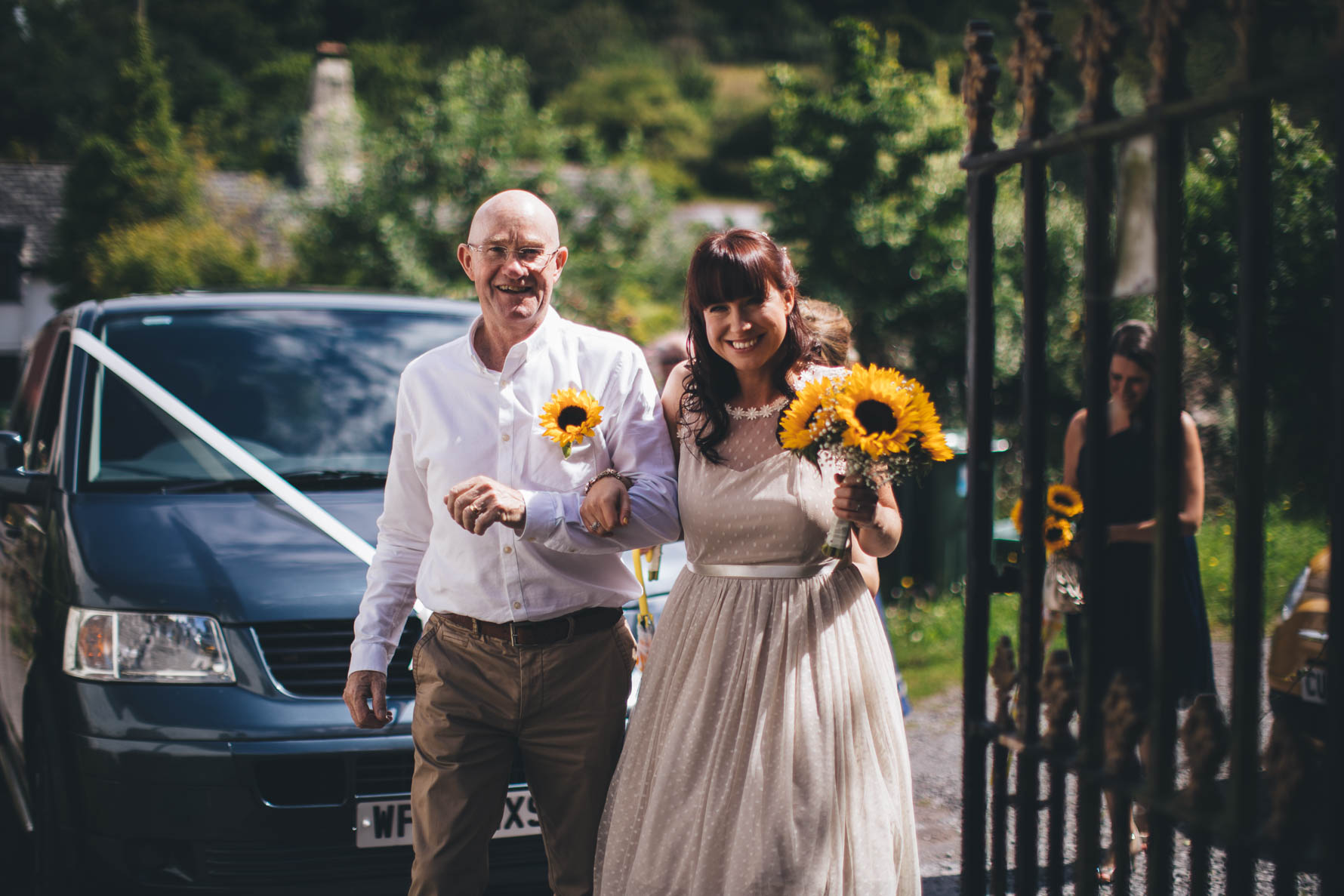 Bride and her father walking into a church. The bride is holding a bouquet of sunflowers and her father is wearing a white shirt and khaki chinos with a sunflower buttonhole. There is a blue car with a white ribbon on the front behind the two of them and a large iron gate to the right of the picture