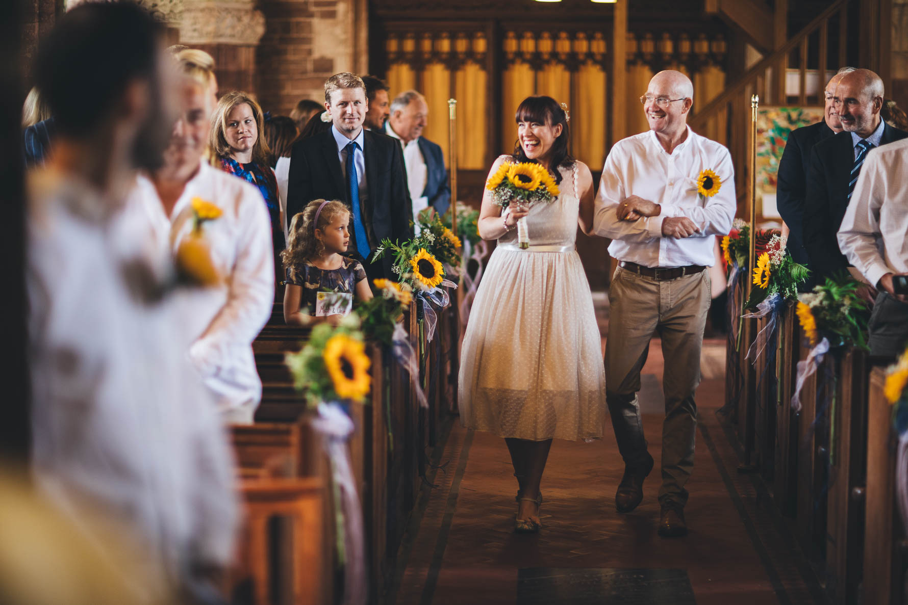 Bride and her father walking down the aisle in a church in Devon. The bride is holding a bouquet of sunflowwers and there are sunflowers on the end of each church pew