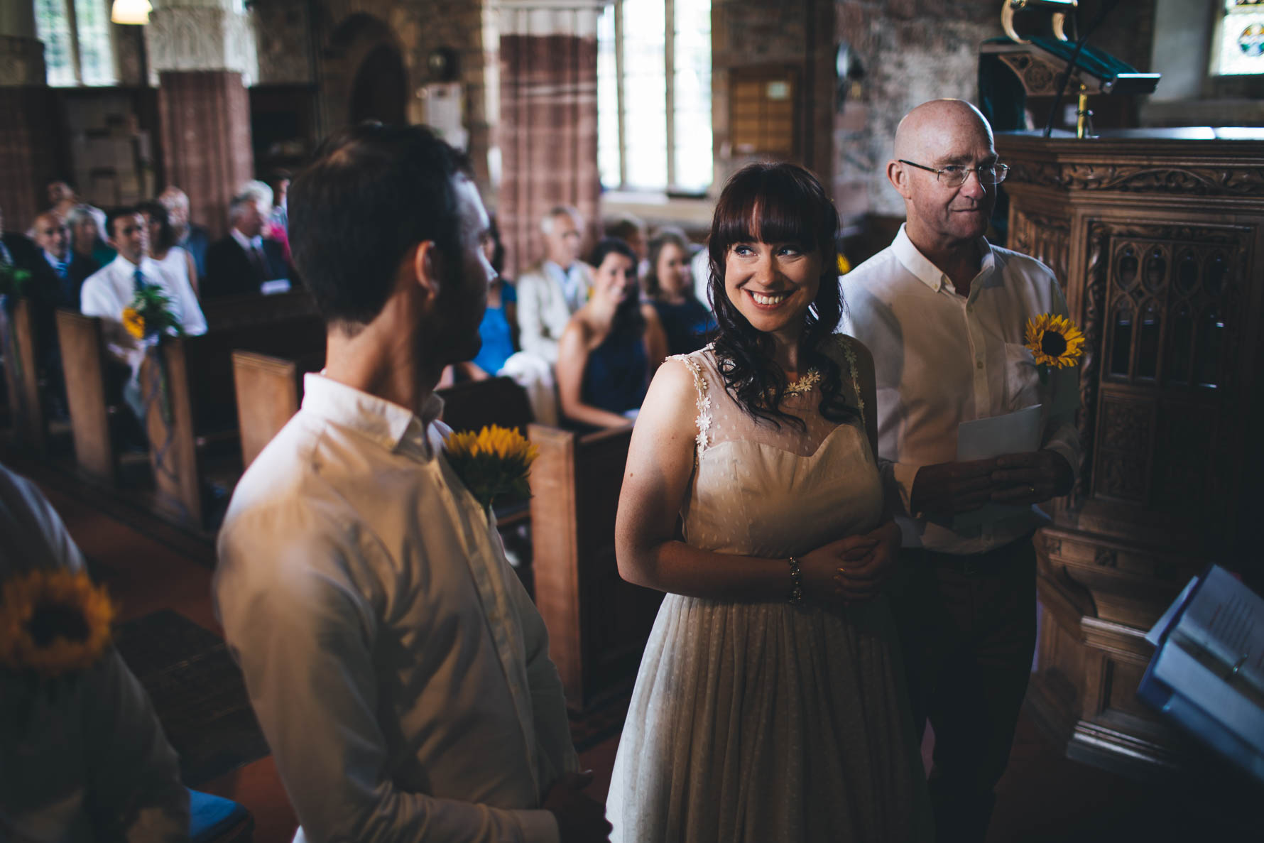Bride looking at the groom and smiling at the front of a church during their wedding ceremony. The bride's father is stood next to the bride.