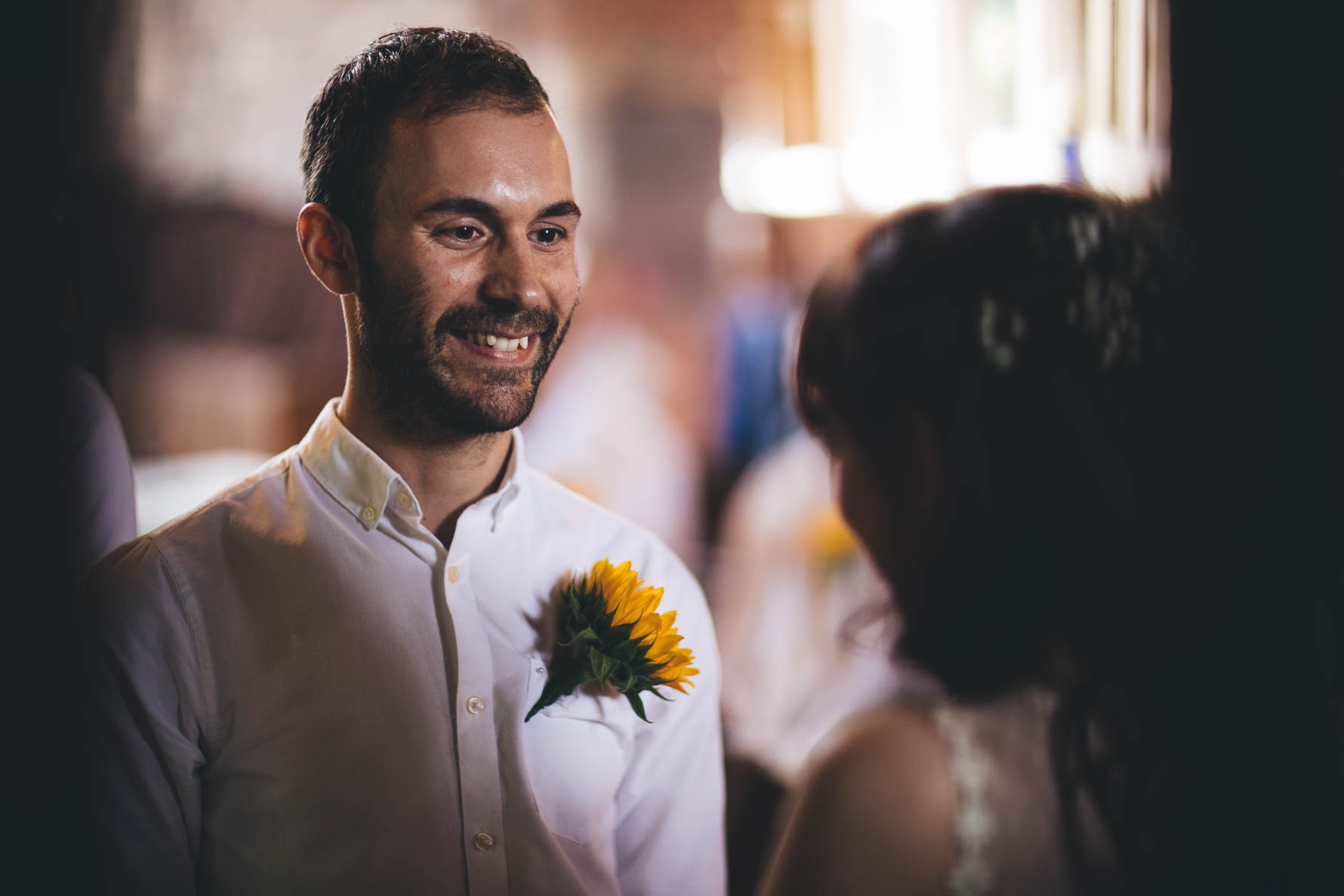 The groom smiling at the bride who is facing away from the camera. The groom is wearing a white shirt and has a sunflower pinned to his shirt