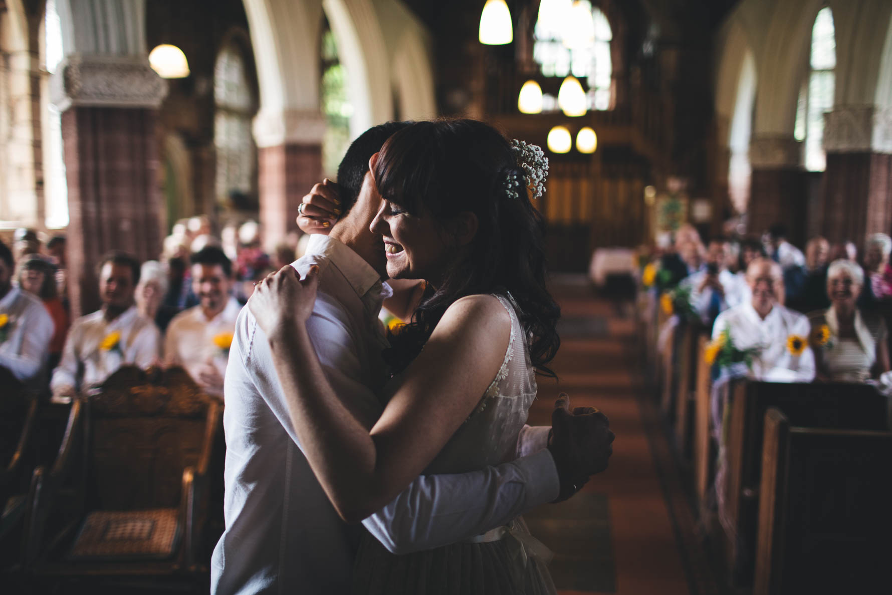 Bride and groom hugging each other at the front of a church in Devon after just getting married. The wedding party is seated in the church pews in the background