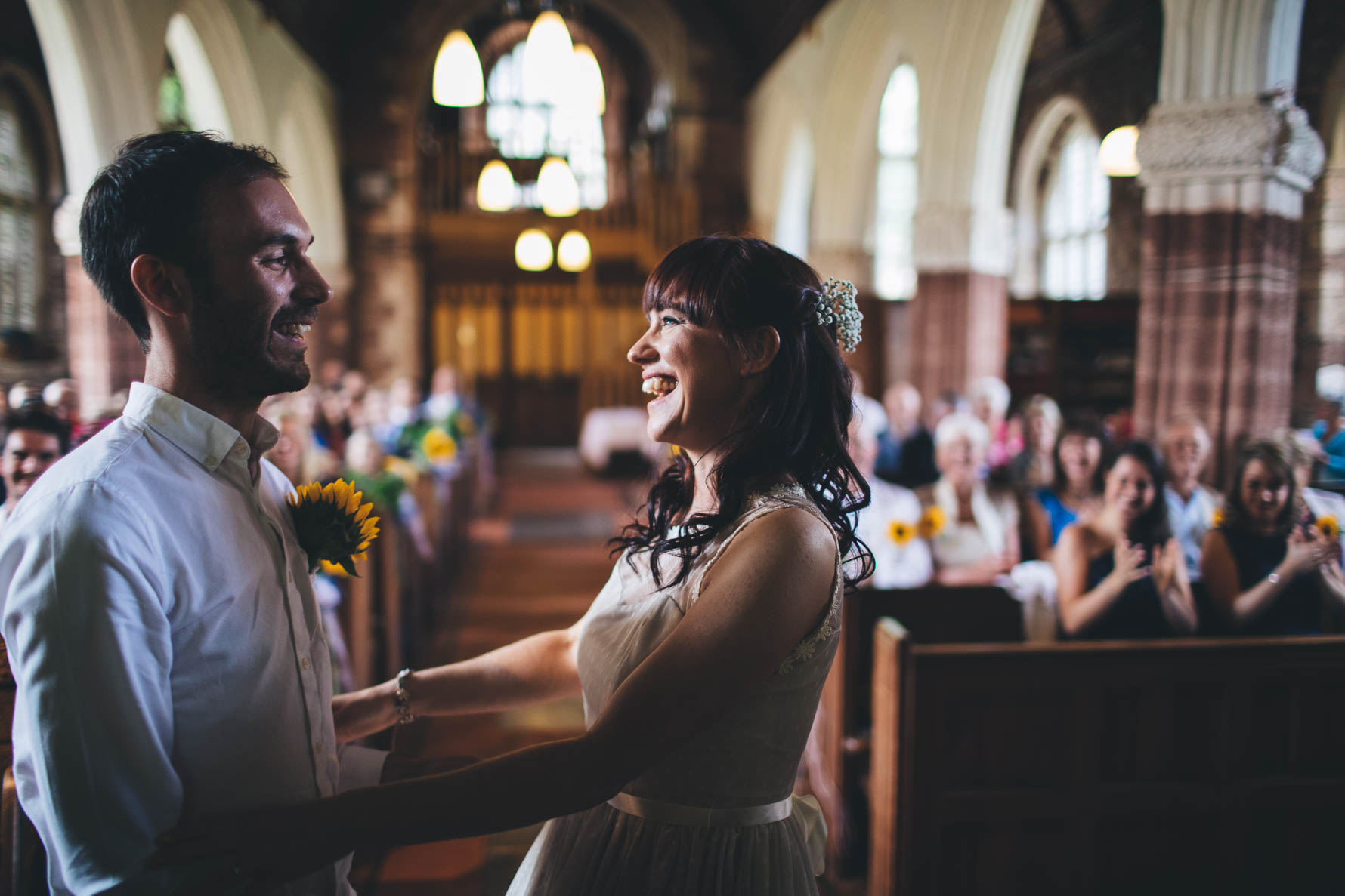 Bride and groom with their arms arund each others waists at the front of a church in Devon after just getting married. The wedding party is seated in the church pews in the background