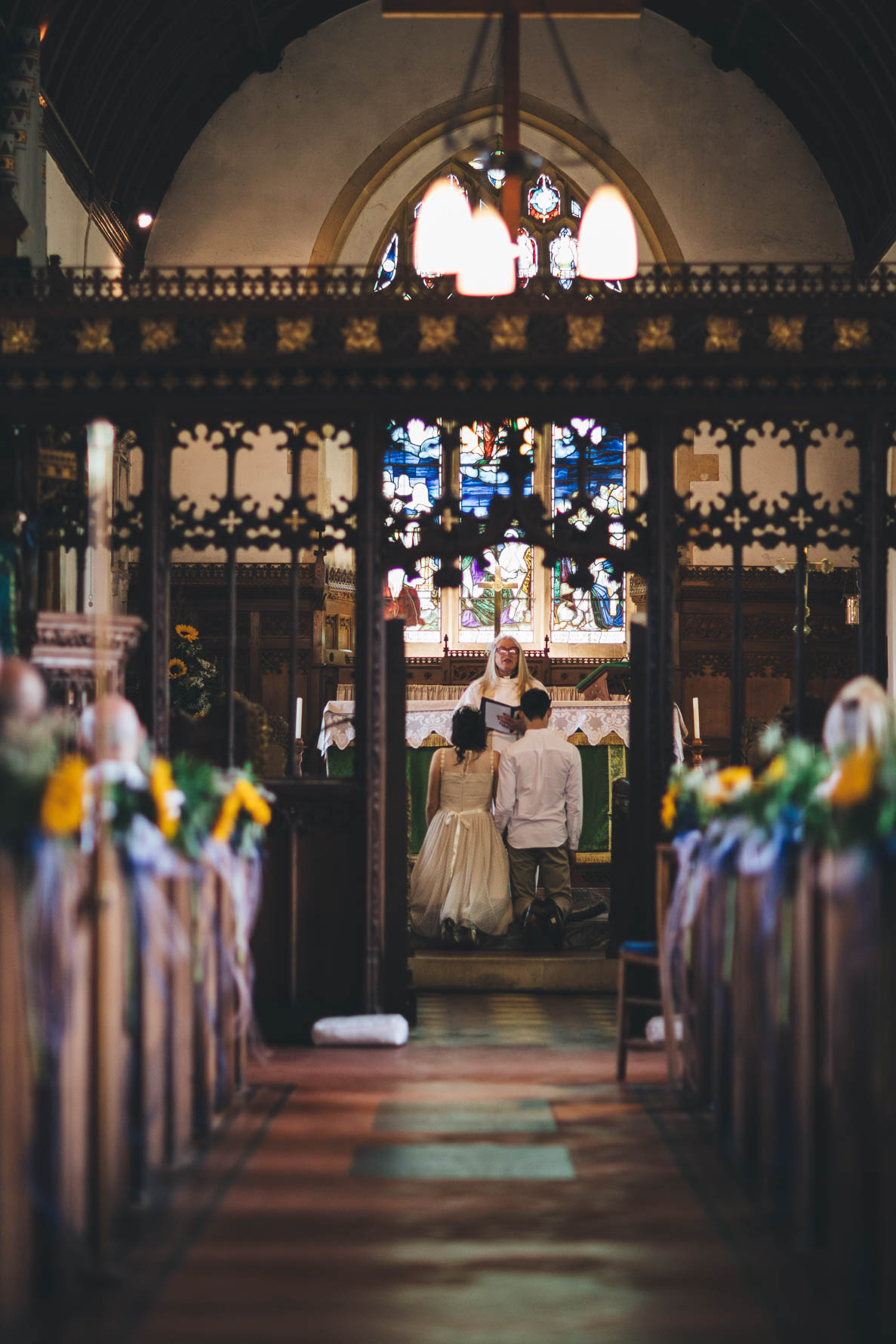 Photo from the end of the aisle looking down the aisle towards the bride and groom who are kneeling in front of a priest during their wedding ceremony. There are large stained glass windos on the back wall above the couple
