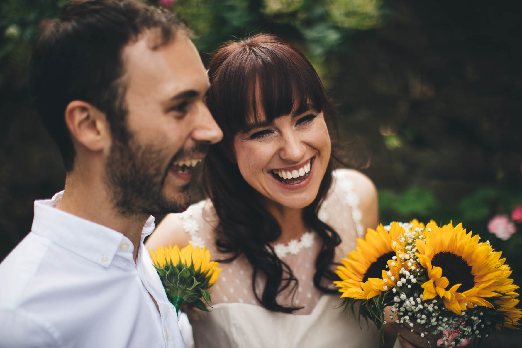 Bride and groom smiling but not looking at the amera. The bride is holding a bouquet of sunflowers and the groom has a sunflower buttonhole on his white shirt