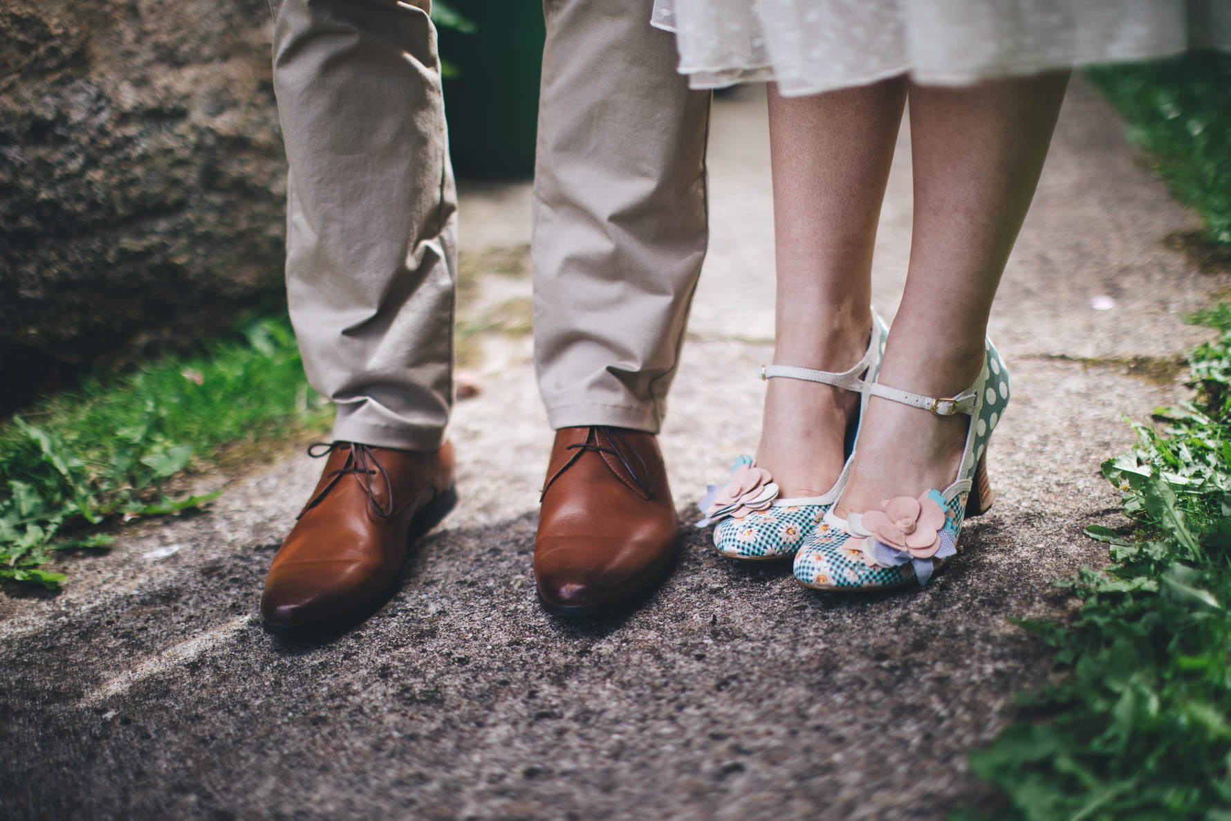 Picture of a bride and groom's shoes on a stone path. The groom is wearing cream coloured chino's and brown shoes and the bride is wearing blue heels with white polkadots and a floral design with a material flower on each shoe