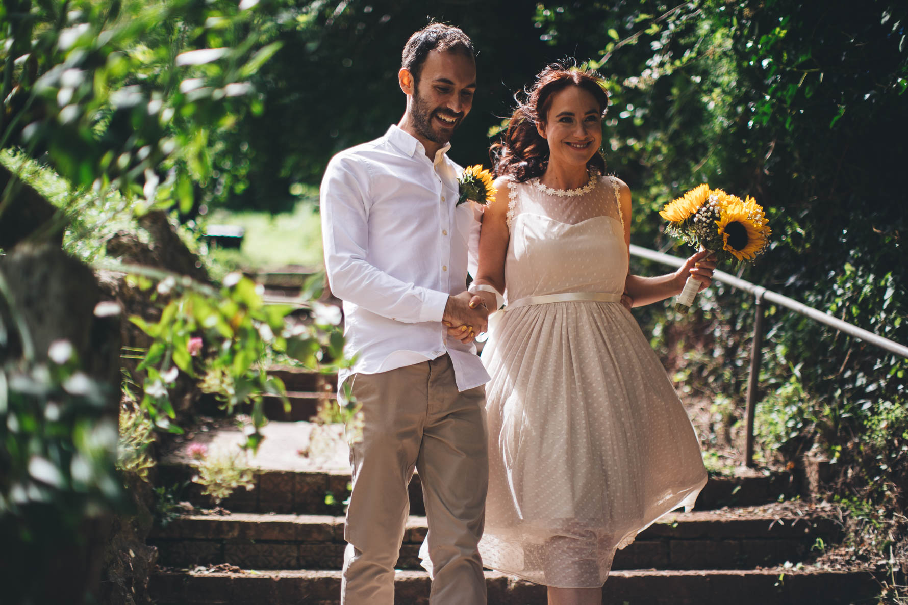 Bride and groom holding hands as the walk down some stone steps in a garden. The bride is holding a bouquet of sunflowers and the groom has a sunflower buttonhole