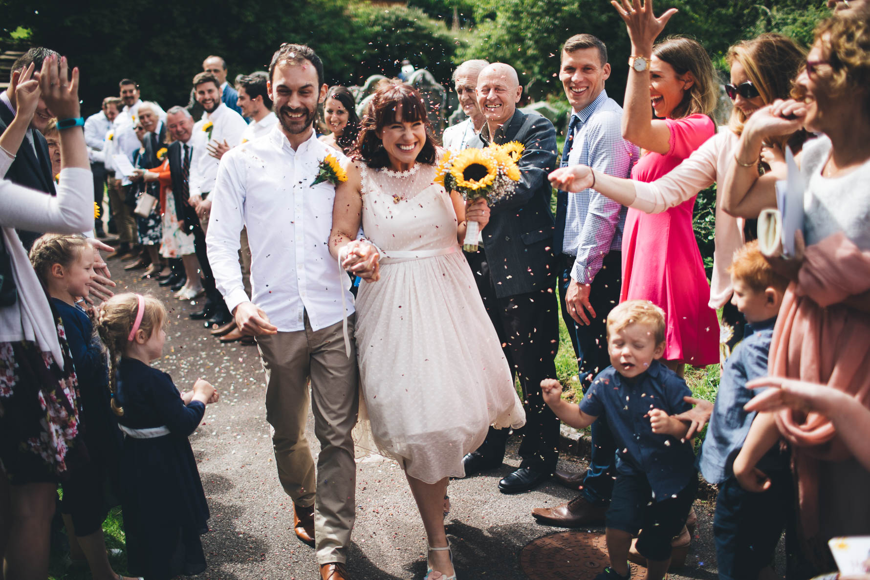 Bride and groom walking down a path with members of the wedding party either side of them throwing confetti over them. The bride is holding a bouquet of sunflowers and both of them have large grins on their faces