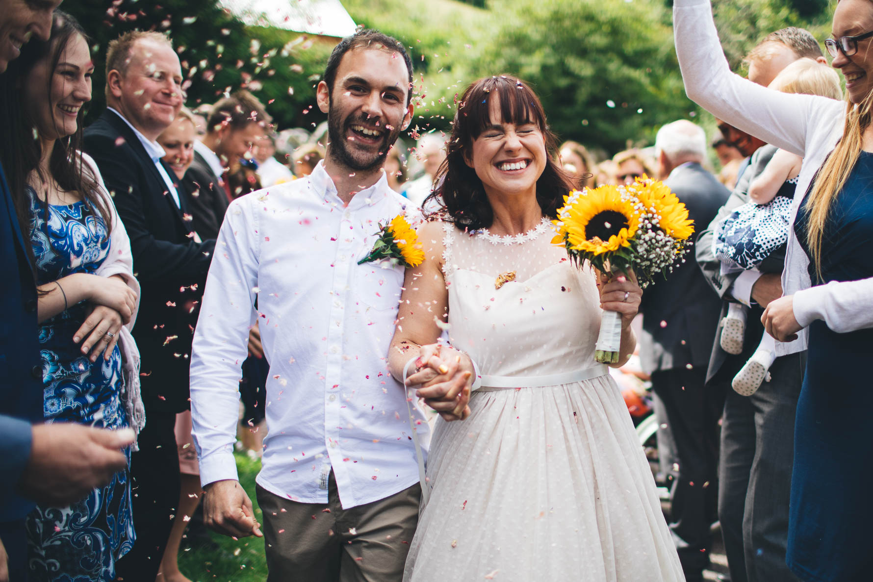 Bride and groom having confetti thrown over them. They are both smiling and the bride has her eyes scrunched up and closed. The bride is holding a bouquet of sunflowers and the groom has a sunflower pinned to his white shirt